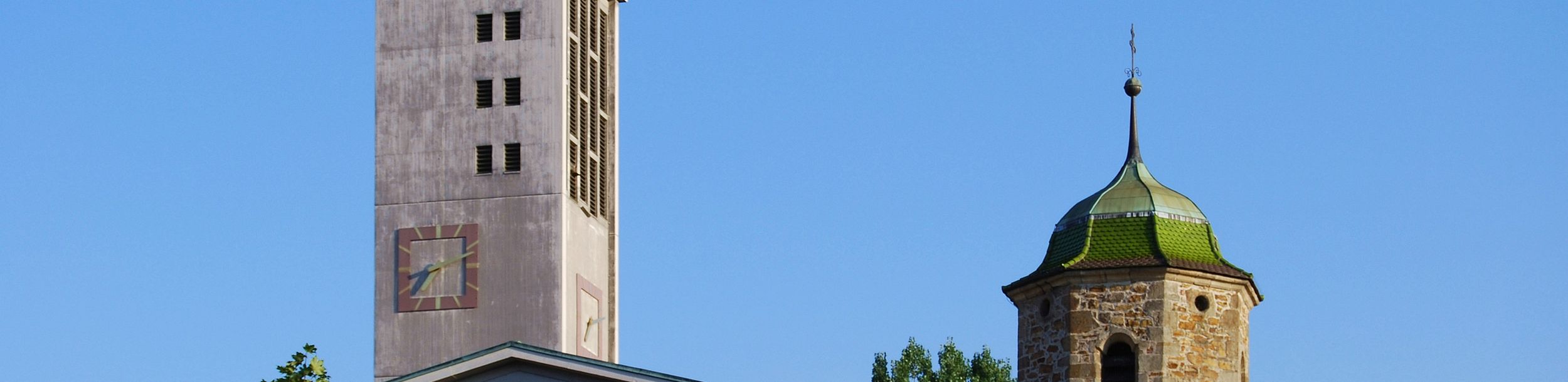 Moderner Kirchturm mit Kreuz neben altem steinernen Turm mit grünem Dach